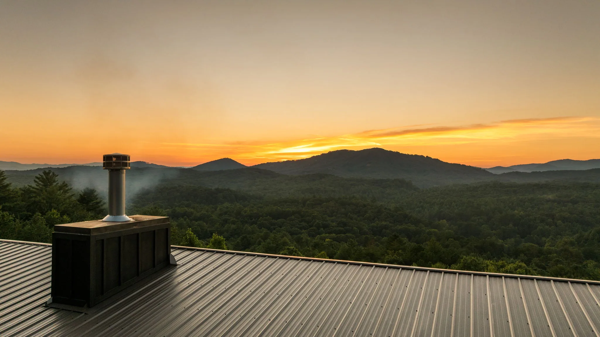 Layered Blue Ridge panorama from the roof line, standing-seam metal and chimney in frame