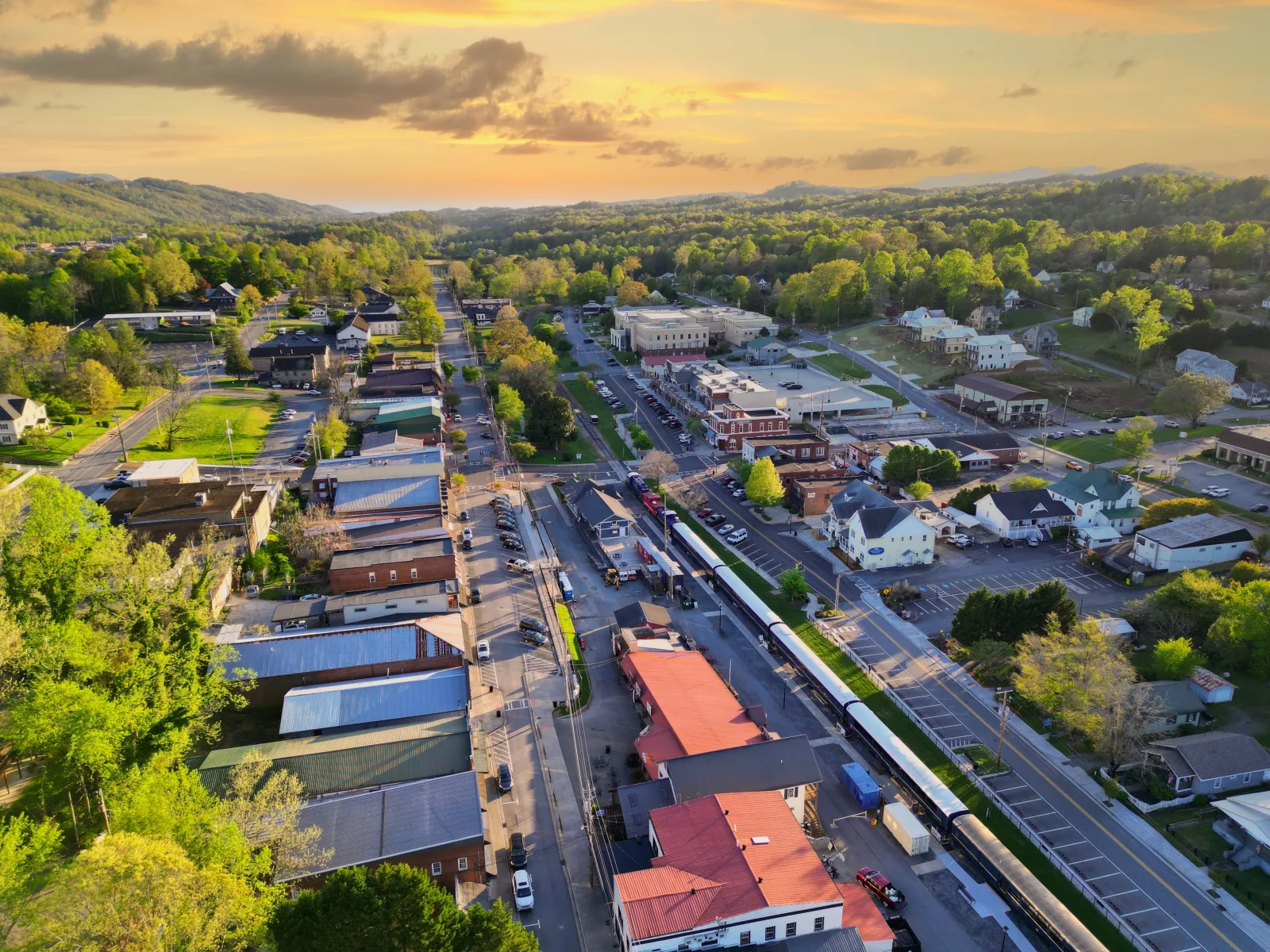 Aerial view of downtown Blue Ridge, Georgia at sunset, with Main Street storefronts, the Scenic Railway train along the tracks, and forested ridges beyond