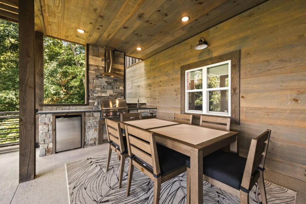 Wide view of the lower patio — stone outdoor kitchen, dining table, and timber ceiling overhead.