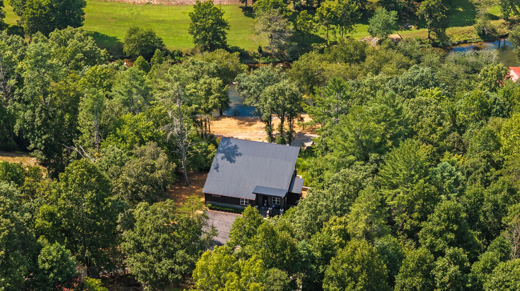 Aerial of the house set into the hardwoods, with the river visible past the ridge.