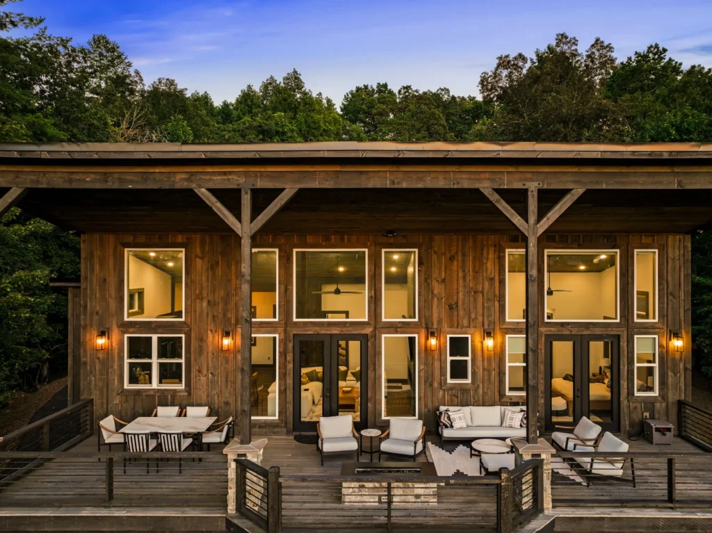 The rear of the house at dusk — the glass wall lit from within and the covered deck furnished and glowing.