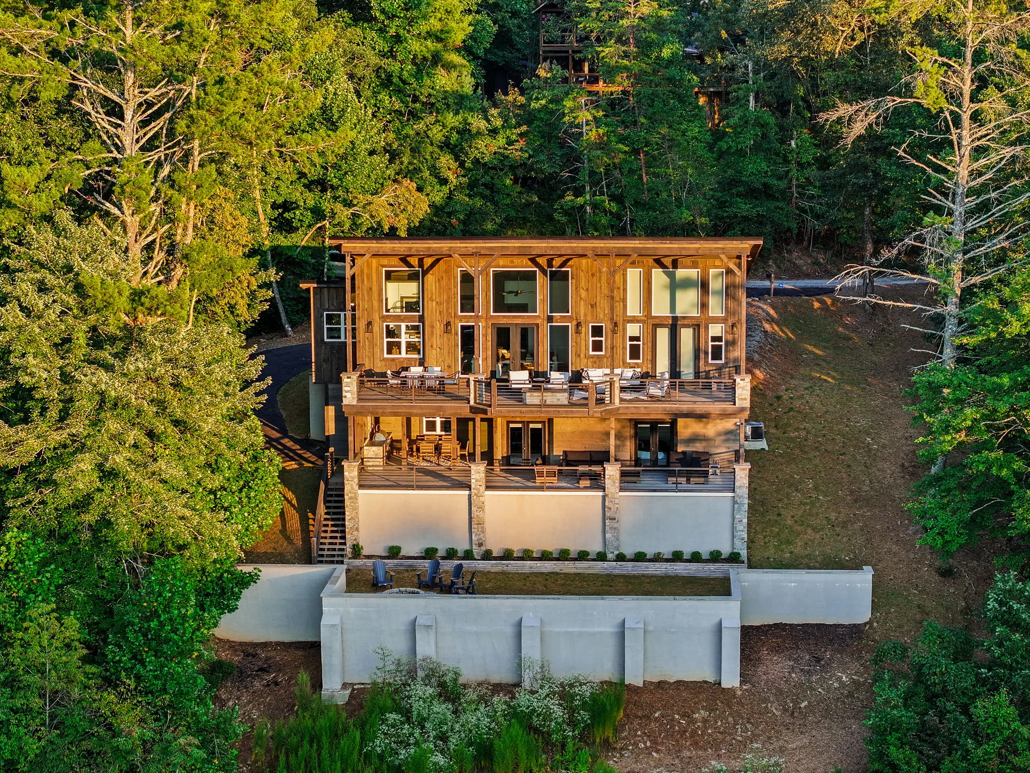 Golden-hour aerial straight onto the rear elevation — two levels of covered deck, the terraced lawn and the fire pit.
